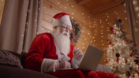 Kamera-Unten-Der-Weihnachtsmann-Sitzt-Zu-Hause-Mit-Einem-Laptop-Auf-Der-Couch-In-Der-Nähe-Des-Weihnachtsbaums-Und-Arbeitet-Mit-Einem-Laptop